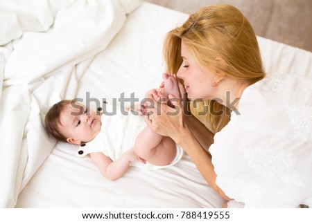 Mother and baby girl on a white bed. Mom and cute baby playing in sunny bedroom at home. Family having fun together. Young mother kissing her daugther feet