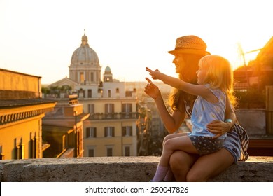 Mother and baby girl sitting on street overlooking rooftops of rome on sunset and pointing
