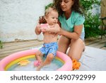 Mother and baby having fun in an inflatable kiddie pool. The baby is holding a toy cup while the mother supports him. Outdoor, summer, happiness, and family time.