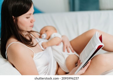 A Mother With A Baby In Her Hands Reading A Book In Bed. Everyday Life Of A Mother With An Infant. Educational Literature And Online Lessons On Newborn Care.