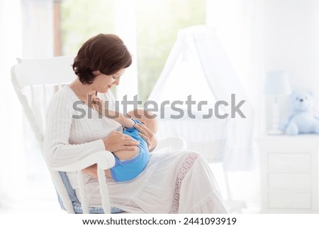 Mother and baby at home. Young woman holding her newborn child sitting in white rocking chair in sunny nursery with crib and garden view window. Parent and kid at home. Infant room interior.