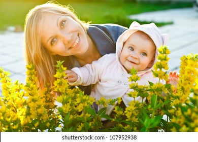 Mother And Baby Looking At Camera Behind Flowers In The Garden With Warm Sunlight