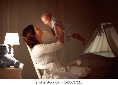 Mother And Baby Read Book In Dark Bedroom. Mom And Child Before Bed Time. Family In The Evening. Kids Room Interior With Night Lamp And Bassinet. Parent Holding Infant Next To Crib.