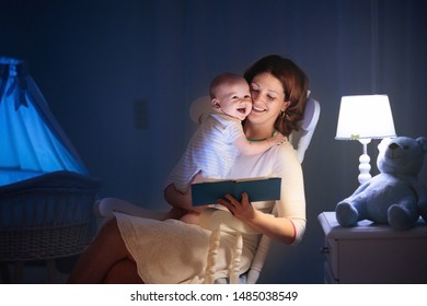 Mother And Baby Read Book In Dark Bedroom. Mom And Child Before Bed Time. Family In The Evening. Kids Room Interior With Night Lamp And Bassinet. Parent Holding Infant Next To Crib.