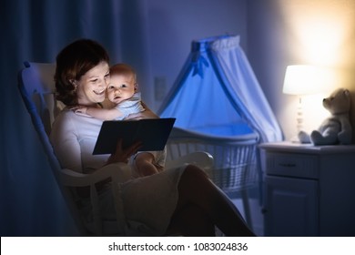 Mother And Baby Reading A Book In Dark Bedroom. Mom And Child Read Books Before Bed Time. Family In The Evening. Kids Room Interior With Night Lamp And Bassinet. Parent Holding Infant Next To Crib.