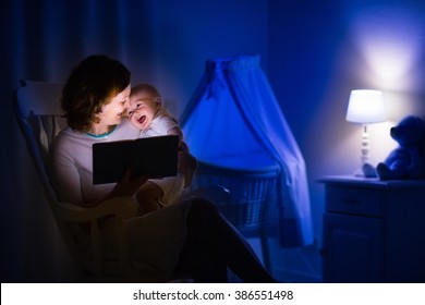 Mother And Baby Reading A Book In Dark Bedroom. Mom And Child Read Books Before Bed Time. Family In The Evening. Kids Room Interior With Night Lamp And Bassinet. Parent Holding Infant Next To Crib.