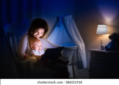 Mother And Baby Reading A Book In Dark Bedroom. Mom And Child Read Books Before Bed Time. Family In The Evening. Kids Room Interior With Night Lamp And Bassinet. Parent Holding Infant Next To Crib.