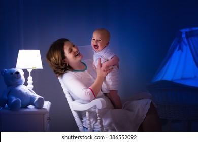 Mother And Baby Reading A Book In Dark Bedroom. Mom And Child Read Books Before Bed Time. Family In The Evening. Kids Room Interior With Night Lamp And Bassinet. Parent Holding Infant Next To Crib.
