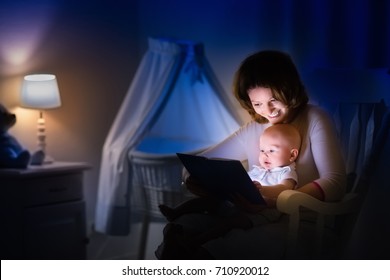 Mother And Baby Reading A Book In Dark Bedroom. Mom And Child Read Books Before Bed Time. Family In The Evening. Kids Room Interior With Night Lamp And Bassinet. Parent Holding Infant Next To Crib.