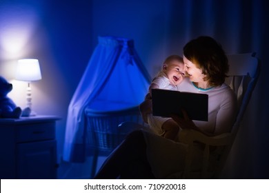 Mother And Baby Reading A Book In Dark Bedroom. Mom And Child Read Books Before Bed Time. Family In The Evening. Kids Room Interior With Night Lamp And Bassinet. Parent Holding Infant Next To Crib.