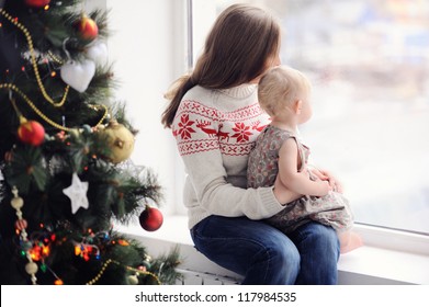 Mother With Baby Sitting By The Window. Christmas Decorations. On Christmas, Waiting For A Miracle. A Woman With A Child Looking Out The Window.