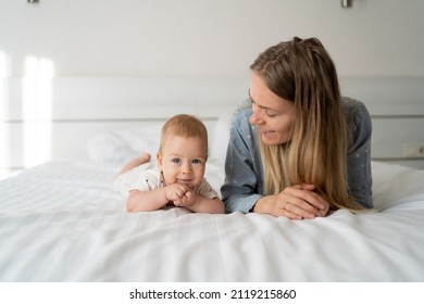 Mother With Baby Six Months Old In The Bedroom. Young Happy Mum Lies Next To Her Adorable Baby Daughter Playing In White Bed Enjoying Sunny Morning In Modern Home Bedroom