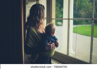 A mother with a baby in a sling is standing by the window in a grand country house