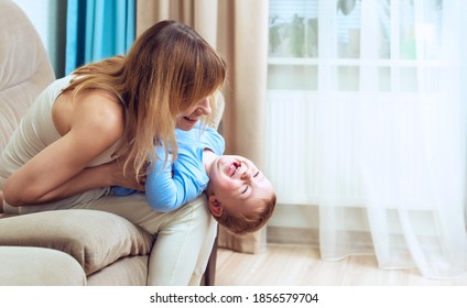 Mother And Baby Son Plays Tickle At Home On The Sofa Living Room. Happy Motherhood And Childhood