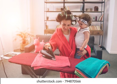 Mother And Baby Together Engaged In Housework Ironing Clothes . Housewife And Kid Doing Homework. Woman With Little Child In The Living Room. Homemaker Doing Many Tasks While Looks After Her Infant