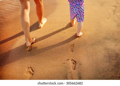 Mother And Baby Walking Along The Beach. Legs, Feet And Footprints On The Sand. Summer Vacation.