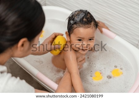 mother bathing her cheerful infant baby with a soft sponge with foam bubbles in a bathtub 