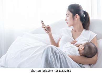 Mother Breastfeeding Her Newborn Baby Beside Window. Milk From Mom’s Breast Is A Natural Medicine For Children. Young Woman Feeding Baby. Mom Using The Phone.