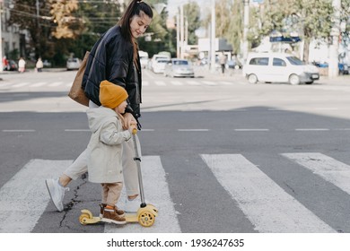 Mother Brings A Child Riding A Scooter On The Crosswalk 