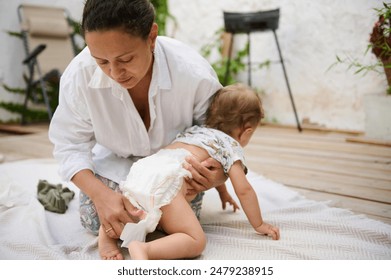 A mother carefully changes her baby's diaper on an outdoor patio, conveying care and bonding in a natural setting. The scene is intimate and captures everyday parenthood moments. - Powered by Shutterstock