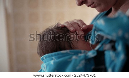 Mother caressing her baby’s head while wearing a baby carrier, capturing a tender and nurturing moment in the kitchen, highlighting the bond and care between mother and child