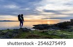 A mother carrying her baby in a backpack stands on the rocky coast of Vancouver Island, Canada, admiring a tranquil sunrise over the ocean. A serene moment of nature and family bond.