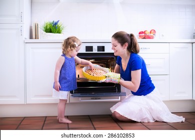 Mother And Child Bake A Pie. Young Woman And Her Daughter Cook In A White Kitchen. Kids Baking Pastry. Children Helping To Make Dinner. Modern Interior With Oven And Other Appliances. Family Eating.