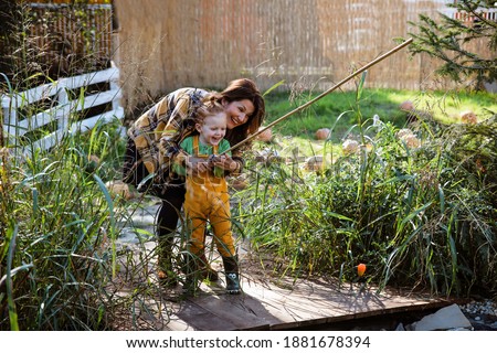 Mother and Child Boy fishing Together, standing on a dock in green grass around. Female Parent teaching son how to Fishing outdoors
