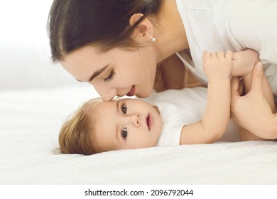 Mother And Child Connection. Portrait Of Happy Young Mother Tenderly Kissing Baby Girl Lying On White Bed At Home. Close Up Side View Of Mom With Baby Playing And Spending Time Together.