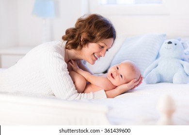 Mother And Child On A White Bed. Mom And Baby Boy In Diaper Playing In Sunny Bedroom. Parent And Little Kid Relaxing At Home. Family Having Fun Together. Bedding And Textile For Infant Nursery.