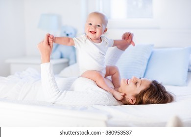 Mother And Child On A White Bed. Mom And Baby Boy In Diaper Playing In Sunny Bedroom. Parent And Little Kid Relaxing At Home. Family Having Fun Together. Bedding And Textile For Infant Nursery.