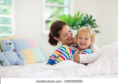 Mother And Child Playing In Bed On Sunny Morning. Mom And Baby Relaxing In White Bedroom At Home. Young Woman Tickling Little Boy. Family Happiness And Love. Parent And Kid After Sleep. Kids Room.