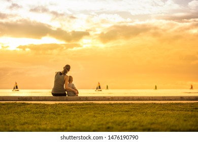 Mother Child Sitting Together Watching Sailboats Float By At Sunset. 