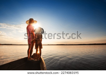 Mother and child swim in boat with seascape in background at sunset. Mother hugs her child. Back view.