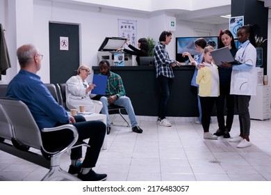 Mother And Child Talking With Pediatrician Holding Laptop While Senior Doctor Shows Form To Patient In Waiting Room. African American Doctor Checking Appointment With Woman And Daughter.