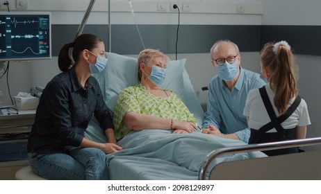 Mother And Child Visiting Retired Woman During Covid 19 Pandemic In Hospital Ward. People With Face Mask Reuniting At Family Visit, Talking To Sick Hospitalized Grandma In Bed.