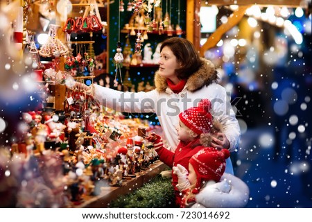 Mother and child in warm hat watching handmade glass Christmas tree ornaments at traditional German Xmas street market. Family with kids shopping for Christmas presents on winter fair on snowy day.
