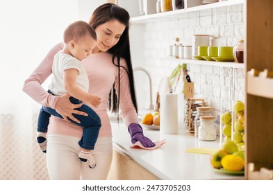 A mother is cleaning the kitchen counter with a cloth while holding her baby. The kitchen has a bright, organized, and clean appearance. Fruit and kitchen utensils are visible on the counter - Powered by Shutterstock