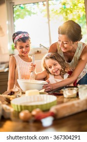 A Mother Is Cooking With Her Two Daughter Of Four And Seven Years Old. They Are Smiling, Wearing Casual Clothes. The Woman Is Helping Her Youngest Mixing The Ingredients In A Large Bowl.