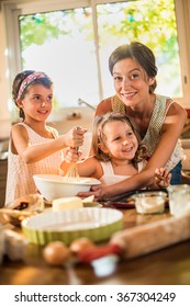 A Mother Is Cooking With Her Two Daughters Of Four And Seven Years Old. They Are Smiling, Wearing Casual Clothes. The Sisters Are Mixing The Ingredients In A Large Bowl With A Whisk.Shot With Flare