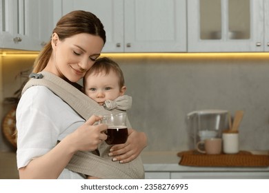 Mother with cup of drink holding her child in sling (baby carrier) in kitchen - Powered by Shutterstock