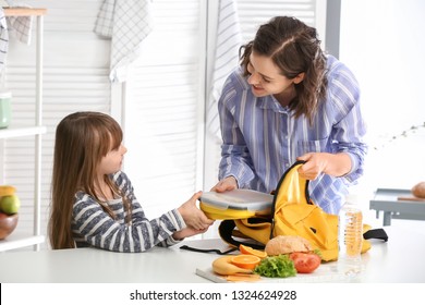 Mother With Cute Daughter Packing Lunch Box Into Backpack At Home