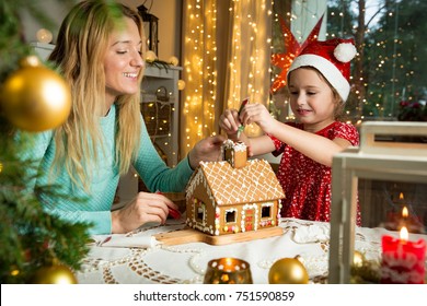 Mother and cute little girl in red hat decorating gingerbread house. Beautiful living room with lights and Christmas tree, table with candles and lanterns. Happy family celebrating holiday together.