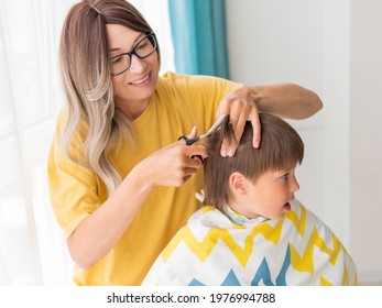 Mother Cuts Her Son's Hair. Little Boy Sits, Covered With Cloth, And Holds Pair Of Scissors. New Normal In Quarantine Of Coronavirus COVID-19. Self Care At Home Lifestyle.