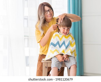 Mother Cuts Her Son's Hair By Herself. Little Boy Sits, Covered With Cloth. New Normal In Case Of Coronavirus COVID-19 Quarantine And Lockdown. Beauty And Self Care At Home Lifestyle.