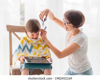 Mother Cuts Her Son's Hair By Herself. Little Boy Sits, Covered With Cloth, And Holds Digital Tablet. New Normal In Case Of Coronavirus COVID-19 Quarantine And Lockdown. Self Care At Home Lifestyle.