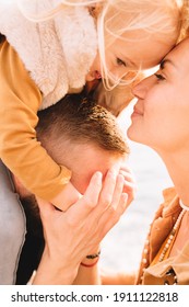 Mother And Dad Kissing Daughter In Her Cheeks. Baby Girl With Blonde Hair, In A Warm Fur Vest. Spring Time, Sun Is Shining. Travel Abroad. Happy Young Family. 