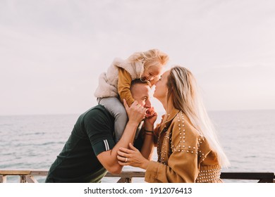 Mother And Dad Kissing Daughter In Her Cheeks. Baby Girl With Blonde Hair, In A Warm Fur Vest. Spring Time, Sun Is Shining. Travel Abroad. Happy Young Family. 