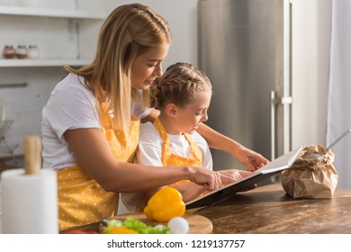 Mother And Daughter In Aprons Reading Cookbook And Cooking Together