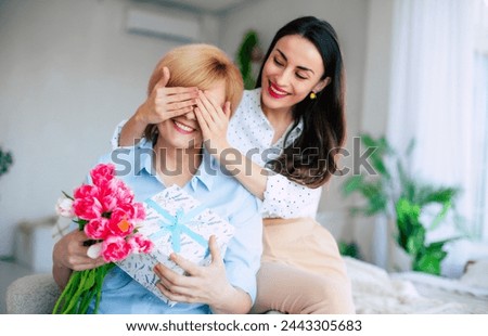 Mother and daughter. Attractive brunette stylish woman covering her mother's eyes while giving her present and gives a bouquet of flowers to tulips. Mothers day, birthday or womens day concept